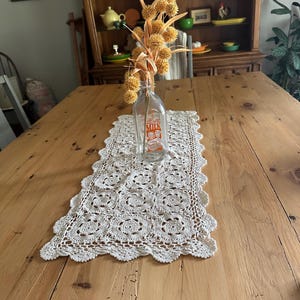 May include: A rectangular off-white crochet table runner with a scalloped edge, featuring a repeating circular floral pattern. A clear glass milk bottle with orange text reading MILK is placed on top, containing a bouquet of dried yellow flowers.