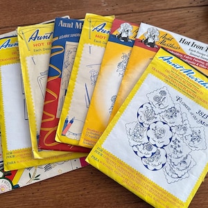 May include: A collection of vintage Aunt Martha's Hot Iron Transfers, featuring various designs. The packets are in yellow, red, and blue, with illustrations and text. One packet is labeled "Flower of the Month 3813".
