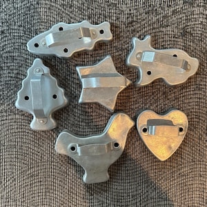 May include: A set of six silver-colored metal cookie cutters in various shapes, including a fish, a rabbit, a Christmas tree, a star, a bird, and a heart. Each cutter features a flat metal handle attached to the top for easy gripping.