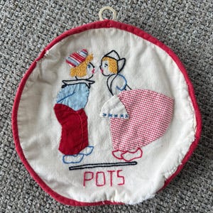 Op de afbeelding: Ronde, witte pannenlap met een rode rand en een ophanglus. Het midden toont een geborduurd ontwerp van twee figuren in traditionele Nederlandse kleding, tegenover elkaar. Het woord "POTS" is eronder in het rood geborduurd.