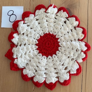 Puede incluir: Un salvamanteles hecho a mano, tejido a ganchillo en forma de flor. El salvamanteles presenta un centro rojo y bordes festoneados rojos, con un patrón blanco en forma de estrella en el medio. Una pequeña etiqueta blanca con el número "8" está en la esquina superior izquierda.