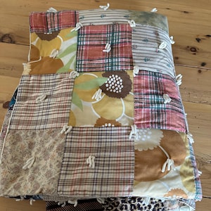 Op de afbeelding: Een patchworkquilt met een verscheidenheid aan stofpatronen, waaronder bloemen-, ruit- en streepontwerpen. De quilt heeft een mix van warme kleuren zoals geel, bruin en rood en is vastgezet met witte garentouwtjes. De onderste laag heeft een luipaardprint.