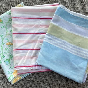 Puede incluir: Tres sábanas dobladas con diferentes patrones. Una sábana tiene un diseño floral con flores rosas y amarillas sobre un fondo azul claro. Otra tiene rayas rosas y rojas. La tercera tiene rayas azules, verdes y blancas.