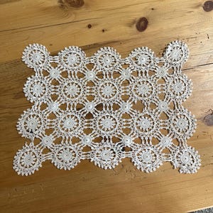May include: A rectangular white doily featuring a repeating pattern of circular crochet motifs connected by intricate threadwork. The design consists of multiple interconnected floral-like medallions with scalloped edges, creating a delicate lace texture.