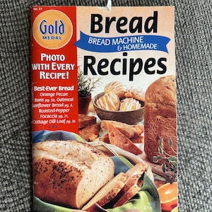 May include: A Gold Medal Bread Machine & Homemade Recipes cookbook featuring a large loaf of bread, a bowl of glazed buns, and slices of bread in a basket. The cover includes text listing recipes like Orange Pecan Buns and Cottage Dill Loaf.