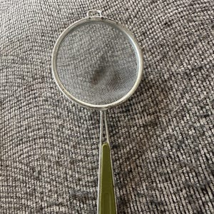 Può includere: Un colino in acciaio inossidabile con un setaccio circolare e un lungo manico. L'impugnatura ha un'impugnatura verde. Questo utensile da cucina è progettato per filtrare liquidi o setacciare ingredienti.