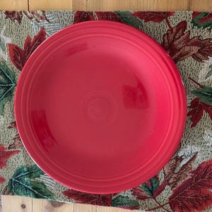 Può includere: Un piatto da cena rosso brillante poggia su un sottobicchiere decorativo con un motivo a foglie autunnali nei toni del verde, rosso e marrone. Il piatto è centrato sul sottobicchiere.