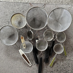 Puede incluir: Colección de coladores de malla de acero inoxidable de varios tamaños, con aros y mangos de metal. Algunos coladores tienen mangos negros o verdes, otros de alambre. Los coladores están dispuestos sobre una superficie gris.