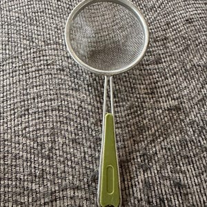 Peut inclure: Une passoire de cuisine dotée d'un bol circulaire en maille d'acier inoxydable et d'un long manche fin avec une poignée en plastique vert clair. Le manche présente une petite découpe rectangulaire au centre et une boucle métallique à l'extrémité.