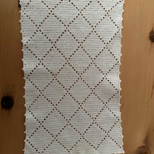 Puede incluir: Un tapete rectangular de ganchillo en color blanco roto que presenta un patrón repetitivo de diamantes creado con puntos calados. Los bordes del tejido están rematados con un delicado borde de picot, aportando un detalle texturizado al diseño.