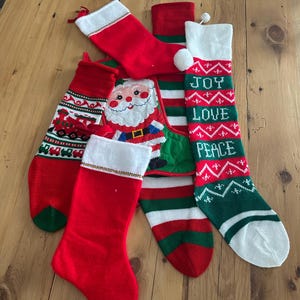 May include: A collection of Christmas stockings in red, green, and white. Some stockings feature Santa Claus, train, and text that reads "JOY LOVE PEACE". The stockings are made of a knitted material.