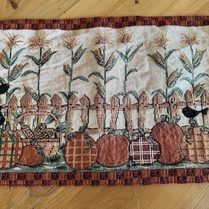 Op de afbeelding: Rechthoekige tapisserie met een herfst oogst thema. Het ontwerp toont maïskolven, een houten hek, pompoenen en merels. De rand is een geruit patroon in tinten rood en bruin. Waarschijnlijk gebruikt als tafelloper of wandkleed.