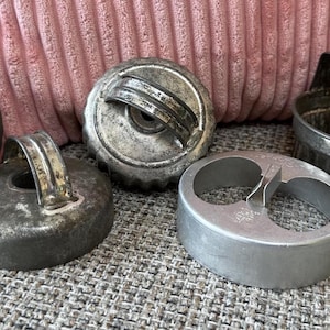 Könnte beinhalten: Eine Sammlung von fünf Küchenwerkzeugen aus Metall, bestehend aus vier runden Keksausstechern mit Griffen und einem ovalen Ausstecher. Die Gegenstände weisen unterschiedliche metallische Oberflächen auf, von verwittertem Dunkelgrau bis zu hellem Silber.