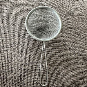 May include: A silver-colored metal kitchen strainer featuring a circular mesh bowl and a long, thin, looped wire handle. The fine mesh screen is framed by a circular metal rim with a small hook at the top for resting on the edge of a container.
