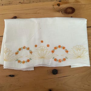 May include: White linen towel with embroidered floral design. The embroidery features orange and yellow flowers and green leaves. The towel is folded on a wooden surface.
