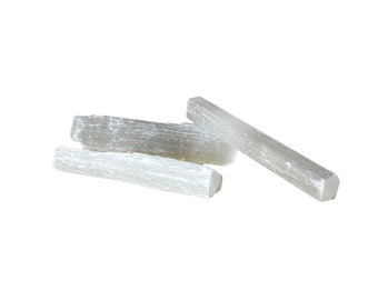 May include: Three selenite sticks, also known as satin spar, are stacked on a white background. The sticks are white and have a striated texture.