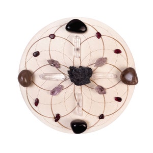 May include: A white circular crystal grid with a black obsidian stone in the center. The grid is surrounded by clear quartz crystals, amethyst crystals, and red garnet stones.