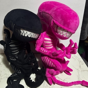 Puede incluir: Dos peluches alienígenas, uno negro y otro rosa brillante. Ambos tienen bocas grandes y abiertas con dientes blancos y cajas torácicas detalladas. Los juguetes son suaves y tiernos, con largas colas y extremidades articuladas.