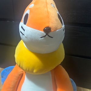 May include: A plush toy featuring an orange and white head with black eyes and a black nose. The toy has a yellow collar and orange body with blue wings. The toy is designed to resemble a cartoon character.