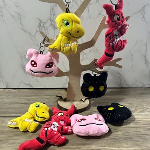 May include: A set of eight plush keychains featuring different characters. The keychains are in various colors, including yellow, pink, red, and black. The characters are all from the Digimon franchise.