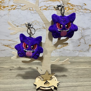 May include: Two plush purple Gengar keyrings with red eyes and pink tongues are displayed on a wooden tree-shaped stand. The keyrings have black metal clasps. The stand has a star-shaped base.