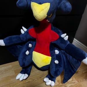 May include: A large plush toy of a Garchomp character, with a navy blue body, yellow face, red chest, and yellow belly. The plush has white claws and accents, with large wings and a detailed design.