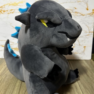 May include: A plush Godzilla toy in a seated position. The toy is primarily gray with blue dorsal fins and accents. It has black claws and a yellow eye detail. The toy is made of soft material and is designed to resemble the iconic movie monster.
