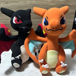 Könnte beinhalten: Zwei Plüschtiere der Pokémon-Charaktere Glurak und Dragoran. Glurak ist orange mit blauen Flügeln und Dragoran ist schwarz mit roten Flügeln.