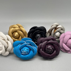 May include: Seven faux leather flower brooches in various colors, including white, gold, black, blue, purple, and pink.