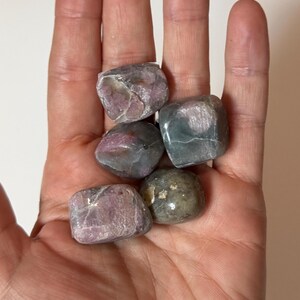 May include: A hand holding five tumbled stones. The stones are a mix of colors, including pink, gray, and green. They have a smooth, rounded shape and appear to be polished. The stones are of varying sizes and shapes.