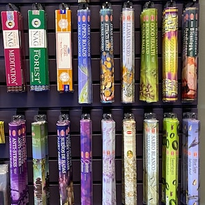May include: A wall display of various incense sticks in colorful boxes. The boxes feature different scents and designs, including floral, spiritual, and nature-inspired themes. The incense sticks are arranged in rows on a dark wooden shelf.