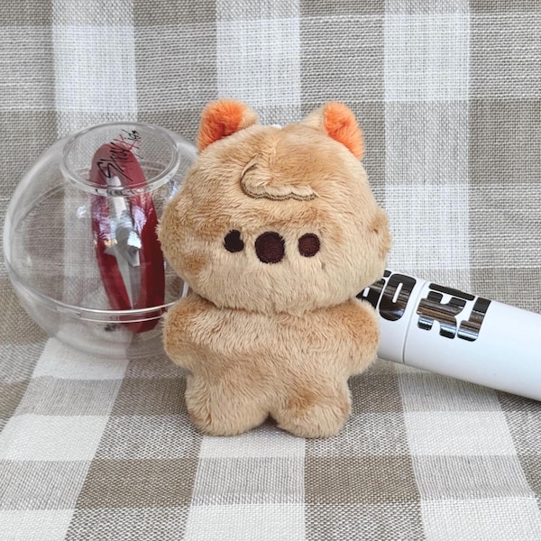 May include: A light brown bear-shaped plush keychain with orange ears and dark brown eyes. A clear glass orb with a red object inside is to the left. A white cylinder with black text is to the right.