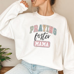 May include: White sweatshirt with the text "Praying foster mama" in colorful block letters with a distressed effect.