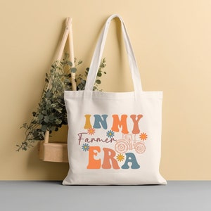 May include: A white canvas tote bag with the text "In My Farmer Era" in colorful retro font. A tractor graphic is included in the design.