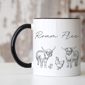 May include: White ceramic mug with a black rim and handle. The mug has a black and white illustration of two Highland cows and two chicks. The text "Roam Free" is written in black cursive on the mug.