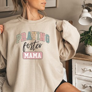 May include: A beige sweatshirt with the text "PRAYING foster MAMA" in colorful letters. The text is in a retro style font.