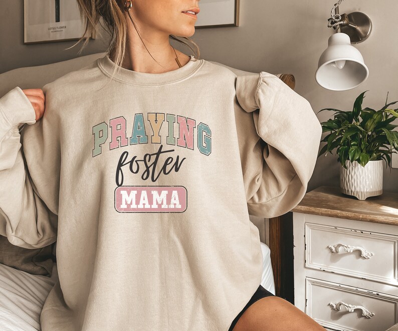 May include: A beige sweatshirt with the text "PRAYING foster MAMA" in colorful letters. The text is in a retro style font.