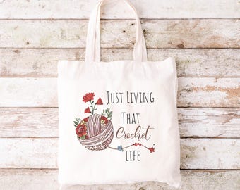 May include: White tote bag with a red, white and green floral design and the text "Just Living That Crochet Life".