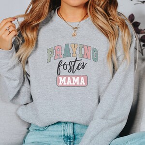 May include: A gray sweatshirt with the text "Praying foster mama" in colorful letters. The text is in a retro style font.
