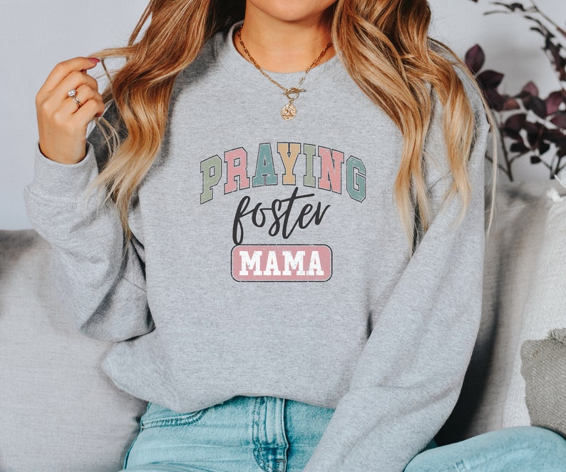 May include: A gray sweatshirt with the text "Praying foster mama" in colorful letters. The text is in a retro style font.