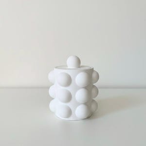 以下が含まれることがあります： 白いセラミック製のジャーで、蓋が付いています。表面は、大きく丸みを帯びた、突き出た球体でテクスチャ加工されています。ジャーの蓋の上には球形のノブが付いています。デザインはミニマリストでモダンです。