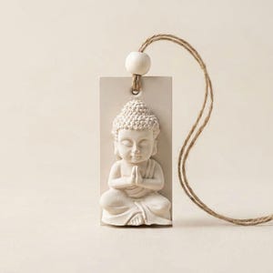 May include: A rectangular off-white hanging ornament featuring a relief of a seated Buddha figure with hands in a prayer position. The ornament includes a single round wooden bead at the top and a natural brown jute twine loop for hanging.