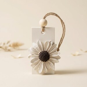 May include: A rectangular white ceramic tag featuring a raised white daisy flower with a dark brown center. The tag has a hole at the top with a natural jute twine loop and a single round wooden bead threaded through the string for hanging.