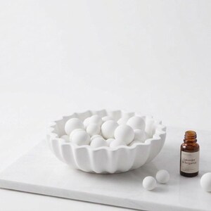 May include: A white scalloped bowl filled with numerous small white spheres, accompanied by a small amber glass bottle labeled Lavender le bergamot and three additional white spheres placed on a white marble surface.