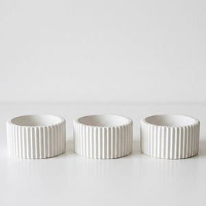 May include: Three white, cylindrical containers with vertical ribbed detailing. The containers are arranged in a row and have a minimalist design. The objects are likely for home decor or storage.