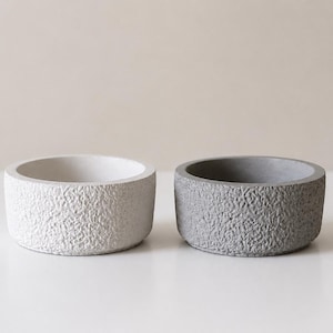 May include: Two small, round, textured concrete bowls. One is light gray, the other is a darker gray. The bowls have a rough, speckled surface and a simple, modern design. They are suitable for small plants or decorative items.