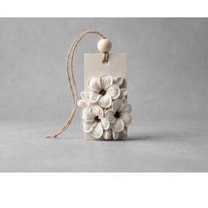 May include: A rectangular off-white ceramic plaque featuring a relief of three white flowers with dark centers. A natural jute twine loop is threaded through a hole at the top, secured by a small, round, light-colored wooden bead.