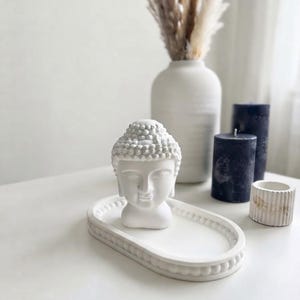 May include: A white Buddha head figurine resting on an oval tray with a beaded rim. The tray and figurine are matte white. Nearby are a tall white vase with dried grasses, two dark blue cylindrical candles, and a small white ribbed container.