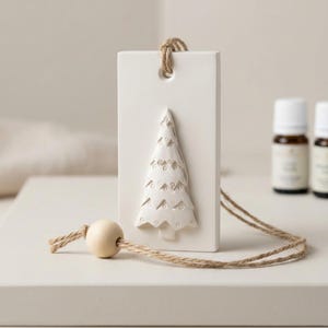 May include: A rectangular white ceramic ornament featuring a raised relief of a Christmas tree. The ornament has a hole at the top threaded with a natural jute twine string and a single smooth wooden bead attached to the hanging cord.