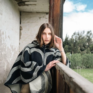 May include: A black and white hooded poncho with a striped design. The poncho is made of a thick material and has a zipper closure. The person is standing on a balcony with a wooden railing, with a view of a green field and blue sky.